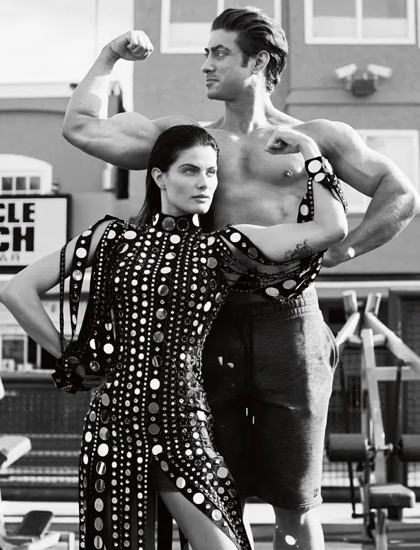 A man flexes his muscles shirtless while a woman in a patterned dress poses beside him. They are outdoors in a gym setting.